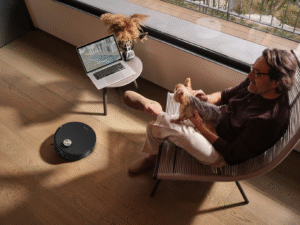 A robot vacuum cleaning a hardwood floor while a man relaxes in a chair with his dog.